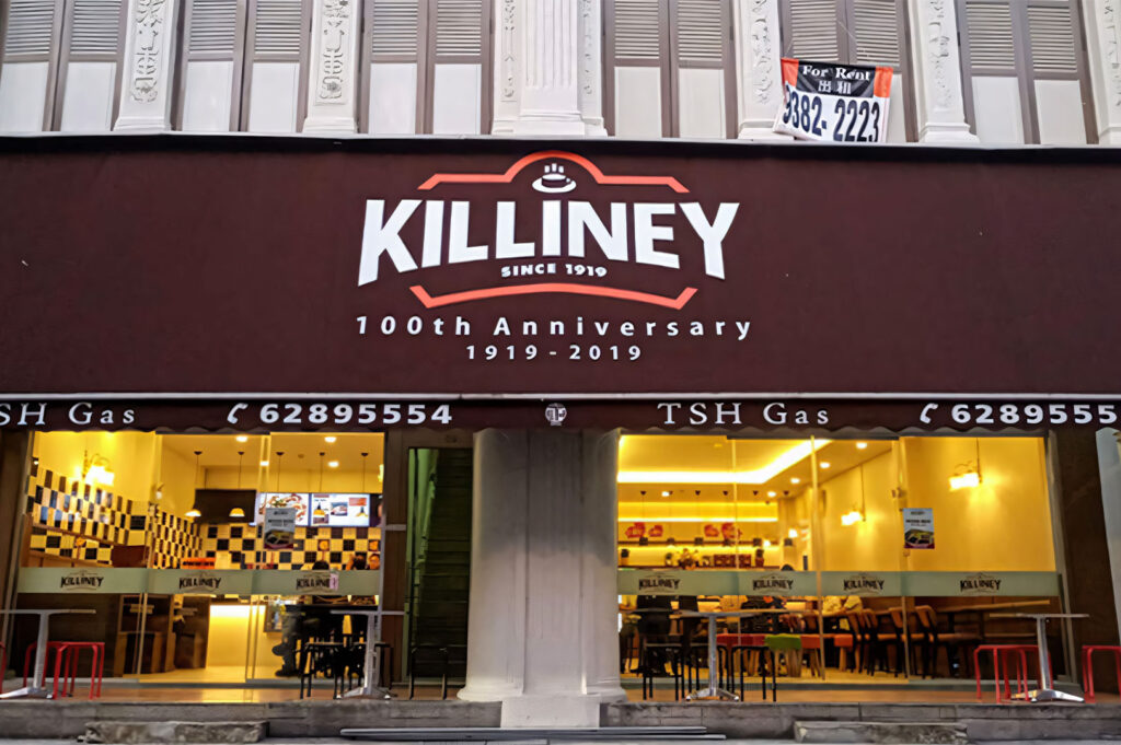 The modern exterior of the Killiney Kopi Tiam coffee shop in Singapore, featuring a large brown sign commemorating its "100th Anniversary" and black and white checkered tiles visible inside.