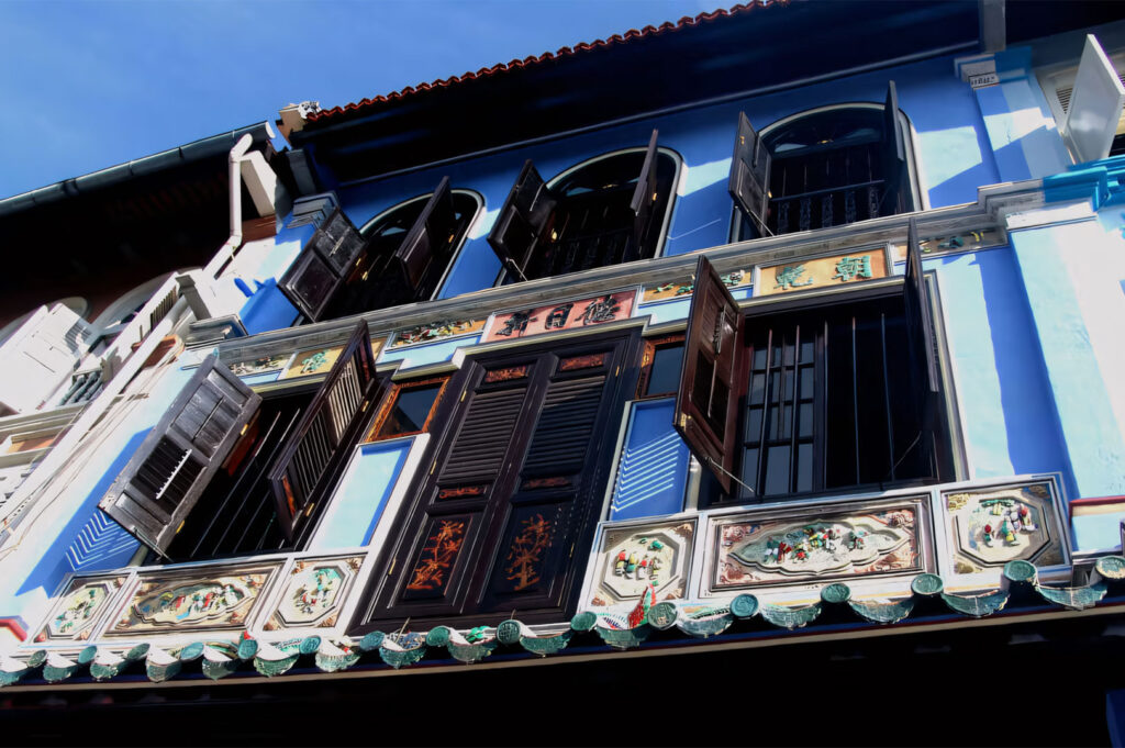 A low-angle close-up of the ornate, bright blue facade of the Baba House in Singapore, highlighting its intricate Peranakan tilework, traditional dark brown wooden shutters, and decorative plaster carvings