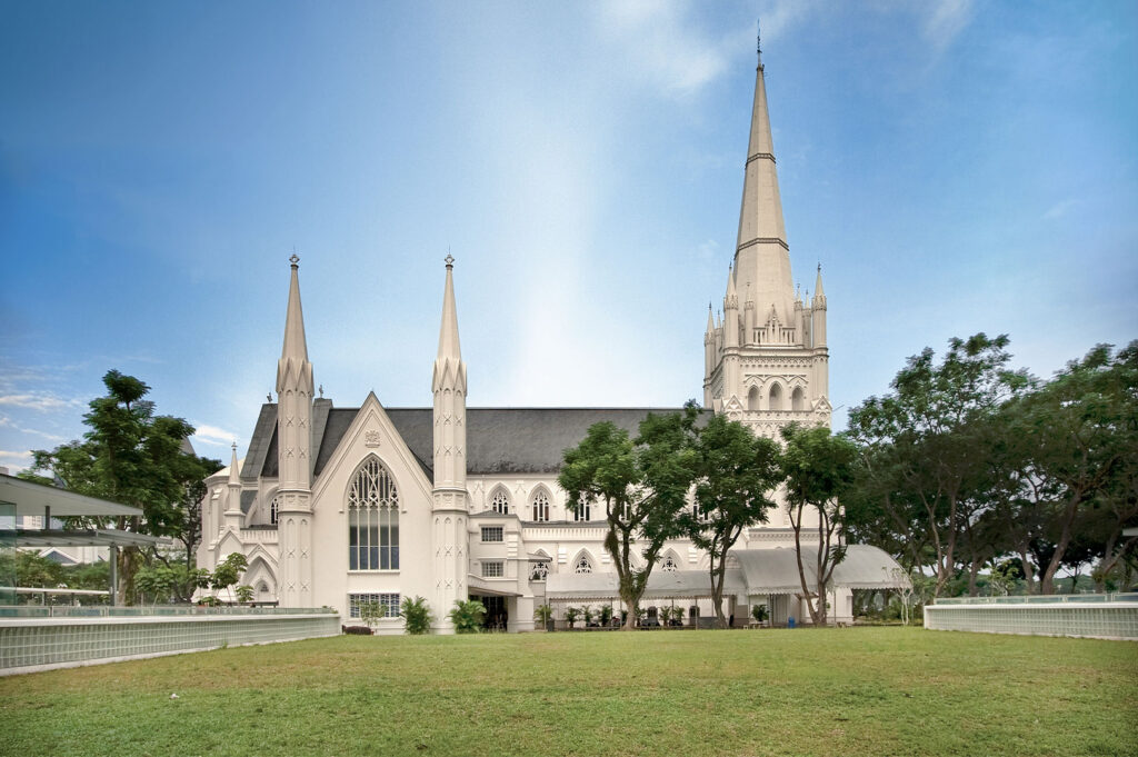 A wide exterior shot showcases St Andrew's Cathedral in Singapore, a magnificent neo-Gothic structure with its prominent spires, against a bright blue sky. The pristine white facade of the cathedral is surrounded by lush green trees and a well-maintained lawn in the foreground. The largest spire stands tall on the right, while two smaller spires are visible on the left. The architectural details, including pointed arches and intricate window designs, are clearly visible, conveying the historical and religious significance of the landmark.