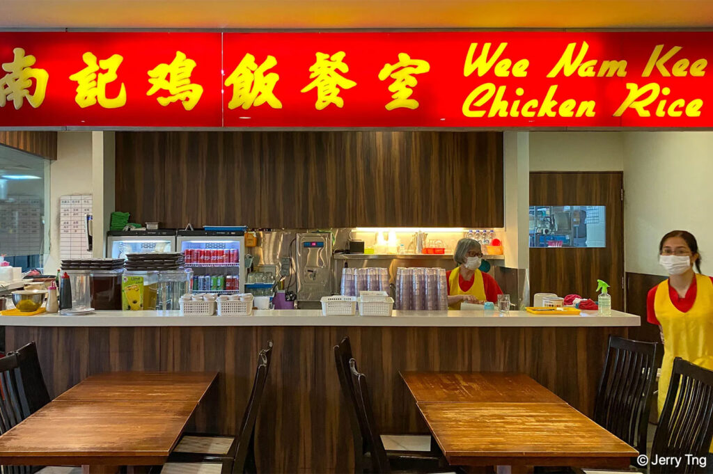 The brightly lit counter of Wee Nam Kee Chicken Rice restaurant, featuring a large red sign with yellow Chinese and English text, tables, and staff in yellow uniforms.