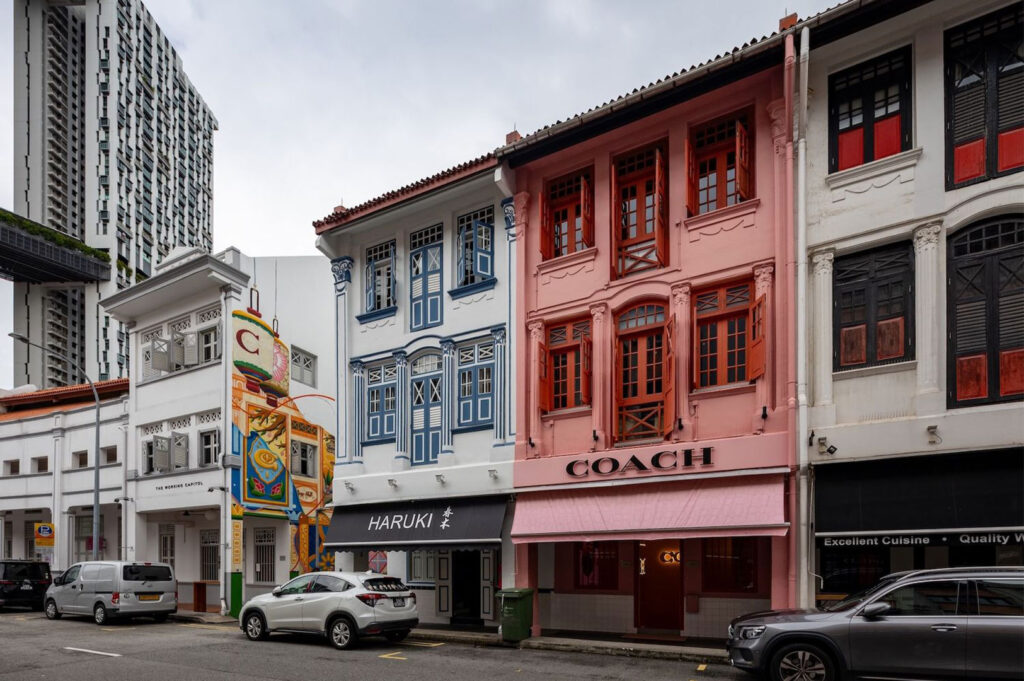 An example of adaptive reuse in Singapore, showing a row of shophouses converted into modern shops, including a pink-painted shophouse occupied by a "COACH" retail store, with a modern skyscraper in the background