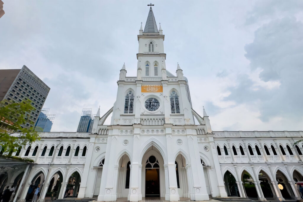 A low-angle shot captures the grandeur of the Chijmes building in Singapore, a former convent school now a lifestyle complex, under a slightly overcast sky. The predominantly white architectural structure, with its Gothic-inspired arches and intricate details, stands prominently against the backdrop of modern skyscrapers visible in the distance. The building's central steeple reaches towards the sky, and numerous windows, some stained glass, adorn its facade. The foreground shows the shaded area beneath the building's arched walkways, hinting at the expansive grounds of the complex.