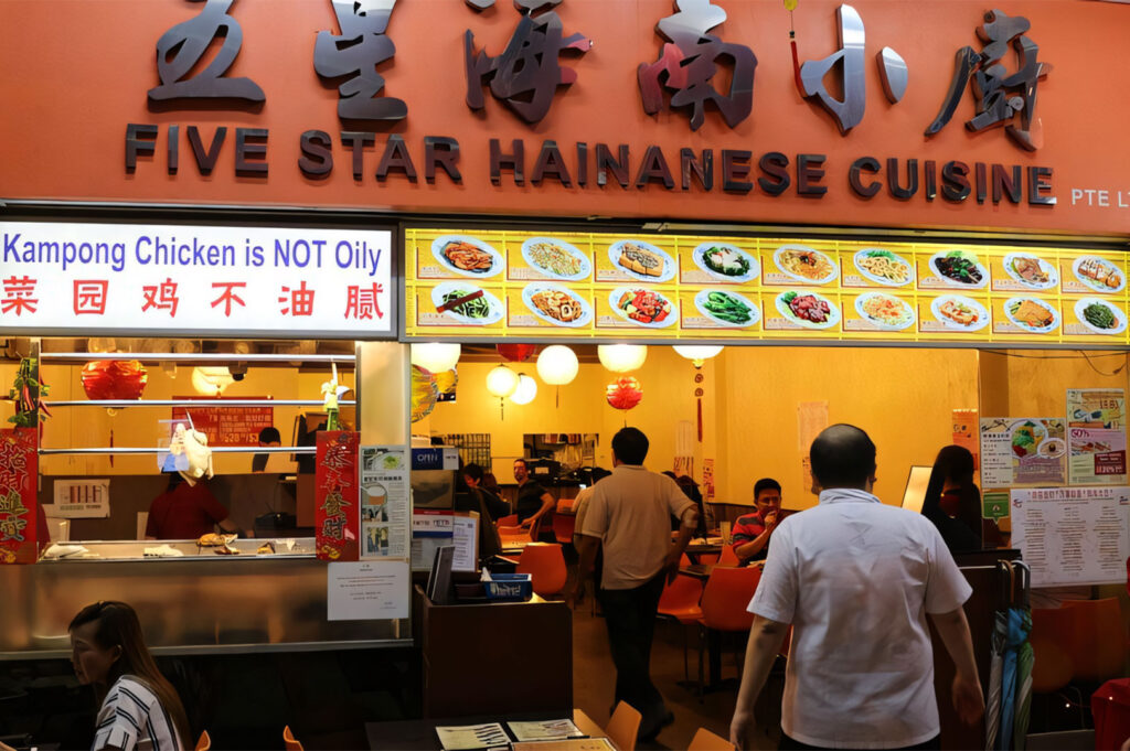 The brightly lit orange entrance and signage for "Five Star Hainanese Cuisine" at a hawker or casual restaurant, showing dining tables and menu boards.