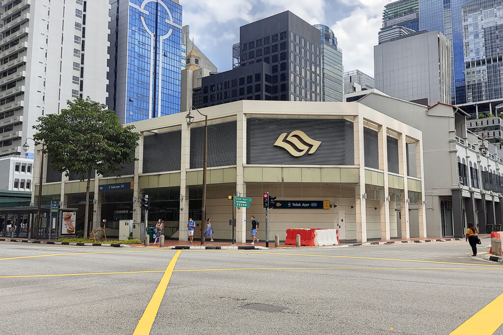 The modern, beige entrance to Telok Ayer MRT Station, located at a busy intersection with surrounding glass skyscrapers of Singapore's Central Business District (CBD).