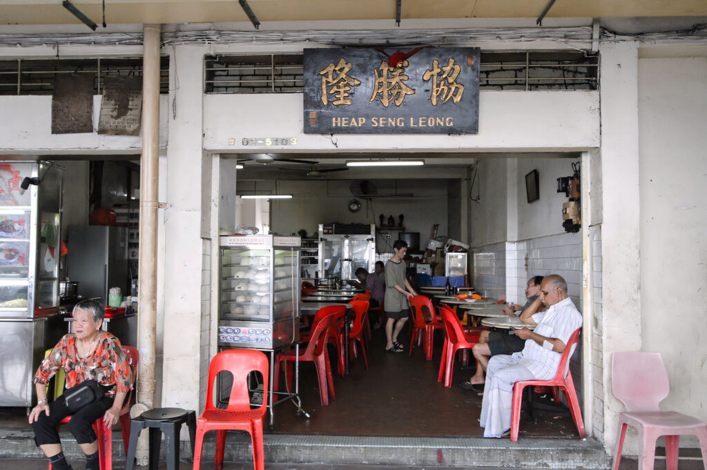 The open shopfront of Heap Seng Leong, a traditional, old-school Kopi Tiam, showing patrons seated on red plastic chairs and a vintage glass display case for steamed buns.