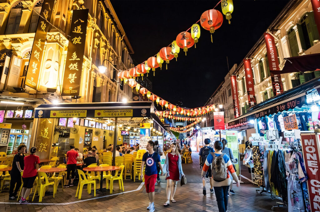 A vibrant night scene on a busy Chinatown street, illuminated by lines of red and yellow lanterns, with people browsing market stalls and dining at outdoor tables.