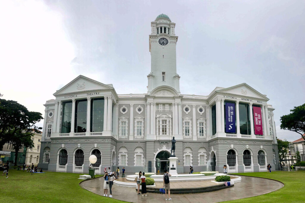 The impressive Victoria Theatre and Concert Hall in Singapore stands prominently under a slightly overcast sky, showcasing its neoclassical architecture. The large white building features a central clock tower, flanked by two symmetrical wings. Banners hang from the right wing, indicating "Victoria Memorial Hall" and advertising a "Home of Theatre" season. A statue stands in front of the main entrance, and a circular fountain is visible in the foreground. People are scattered across the surrounding green lawn and pathways, adding a sense of scale and activity to the historic landmark.