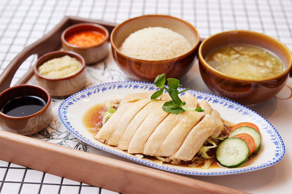 A high-end, elegant serving of chicken rice on a wooden tray, featuring sliced chicken on a decorative platter with cucumber, separate bowls of rice, soup, and three dipping sauces.