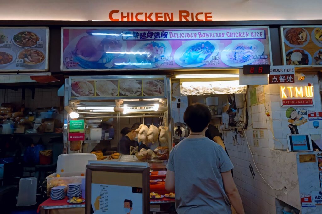 Hawker stall selling delicious boneless chicken rice, featuring overhead menu images of steamed and roasted chicken, hanging cooked chickens, and a customer placing an order.
