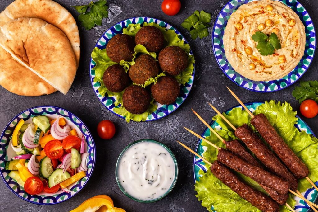 Overhead view of a Middle Eastern meze platter featuring hummus, cucumber salad, minced meat kebabs on skewers, and falafel balls with pita bread.