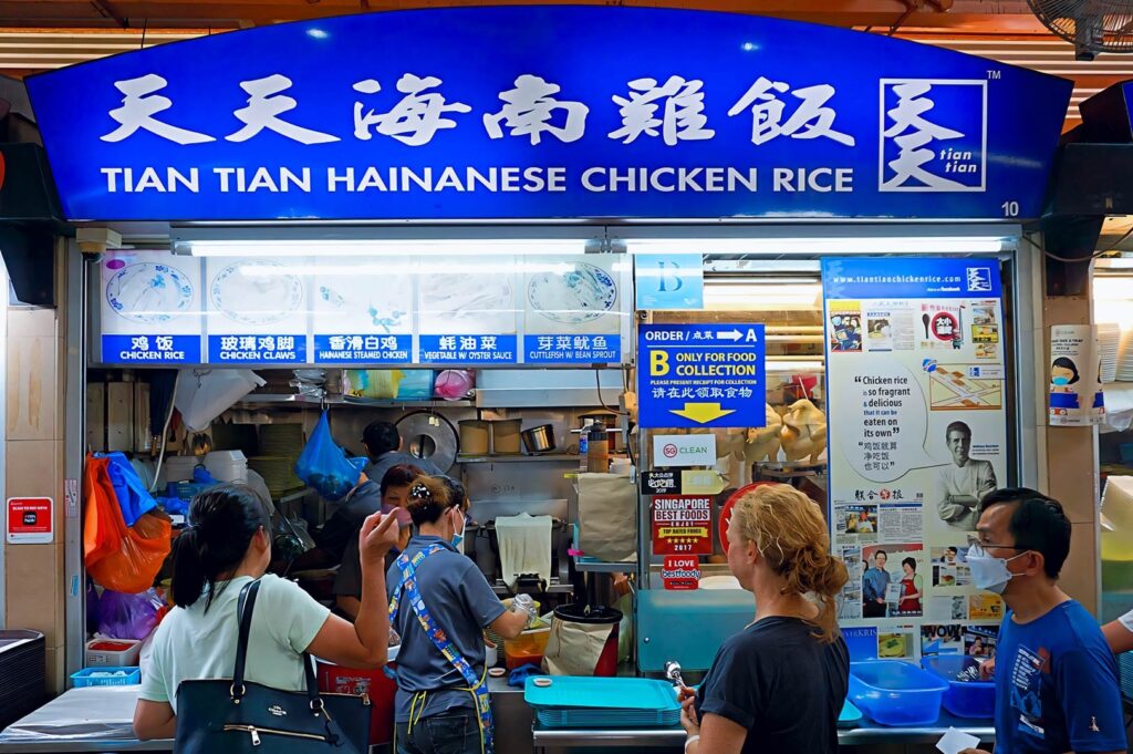 Tian Tian Hainanese Chicken Rice stall with a blue signboard, menu panels showing chicken rice options, and customers queuing at the counter inside a busy hawker centre.