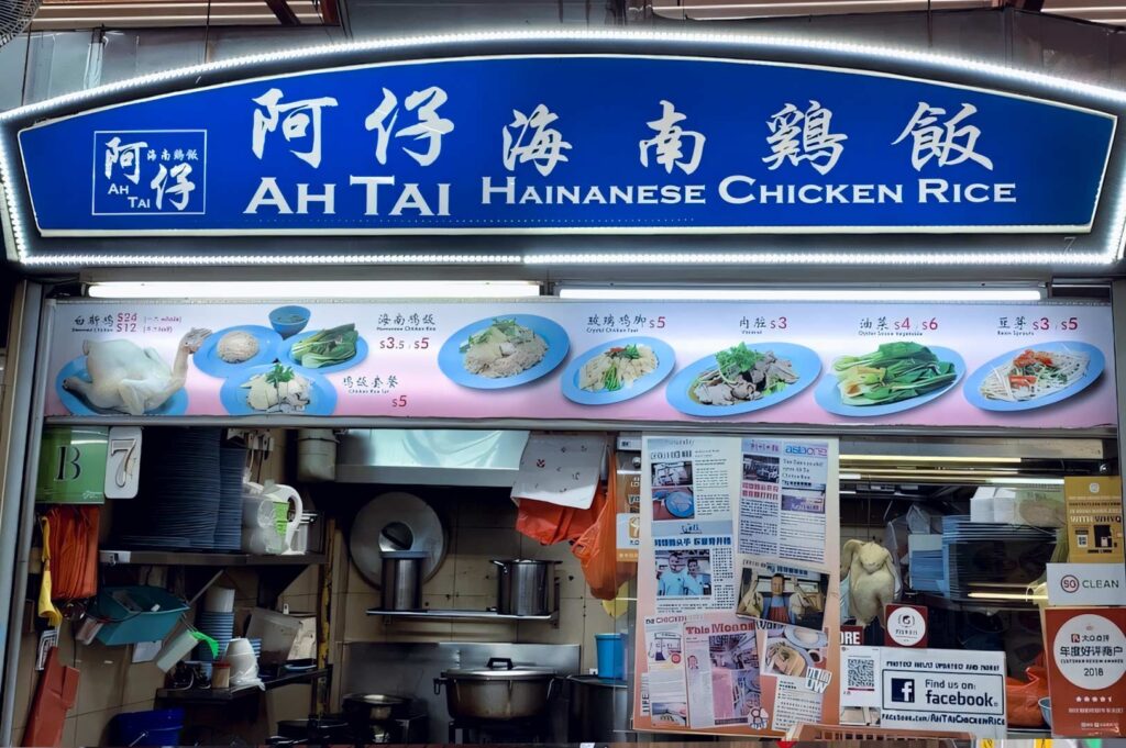 Ah Tai Hainanese Chicken Rice stall with a bright blue signboard, menu photos of chicken dishes, and a traditional hawker kitchen setup filled with pots, utensils, and food preparation equipment.