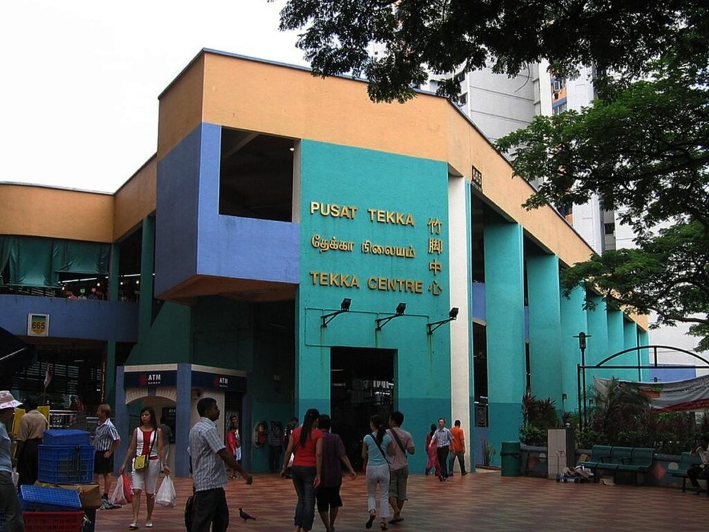 This image shows the exterior of the Tekka Centre in Singapore, featuring a distinctive turquoise and yellow facade with the building's name written in multiple languages. People are seen walking across the paved plaza in front of the entrance, which also houses a small ATM booth and sits under the shade of nearby trees.