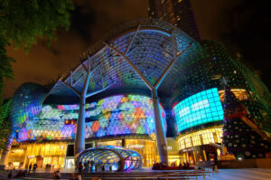 The modern ION Orchard shopping mall facade in Singapore at night, illuminated with vibrant, colorful Christmas light displays and a towering festive tree.