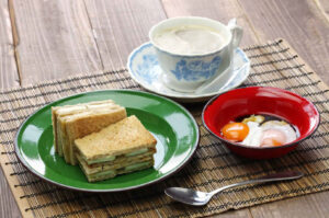 Traditional Singaporean or Malaysian breakfast set: toasted kaya toast slices on a green plate, a cup of kopi (coffee with milk) in a vintage porcelain cup, and soft-boiled eggs in a red bowl.