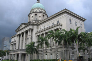 An angled view of the majestic former Old Supreme Court Building (now National Gallery Singapore), showcasing its Neoclassical facade, tall columns, and signature copper dome under a cloudy sky.