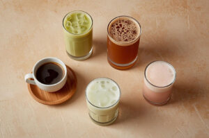 Overhead view of five different cafe beverages: an espresso cup, iced matcha latte, cold brew coffee, iced milk, and a pink latte, arranged on a light background.