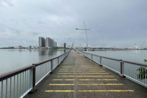 Long, concrete pier walkway extending over the water toward a dense city skyline on a cloudy day, featuring yellow safety lines on the ground.