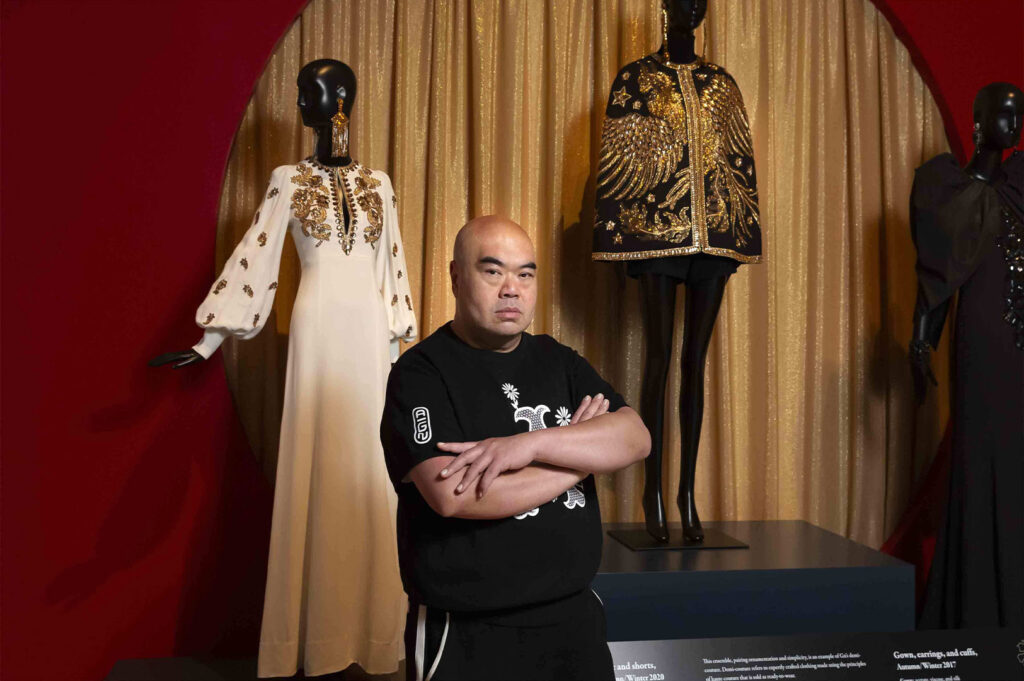 Fashion designer standing with arms crossed in front of three mannequins displaying ornate, historical gowns in a museum or exhibition setting with gold drapery.