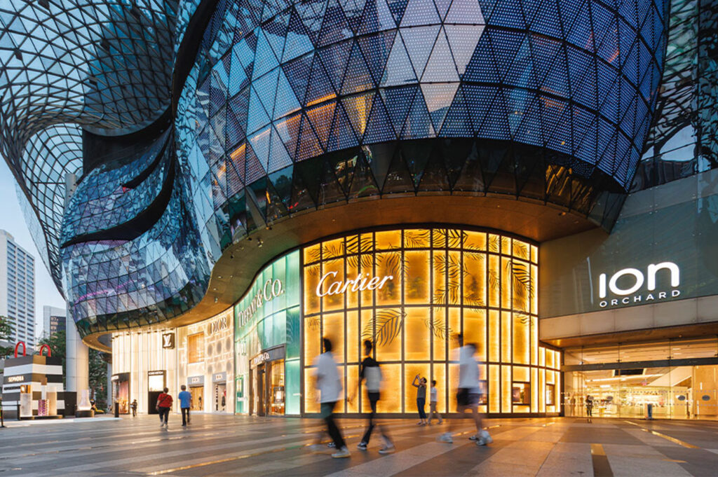 The futuristic, glass-and-steel exterior of the ION Orchard mall entrance on Singapore's Orchard Road during the day, featuring luxury brand storefronts.