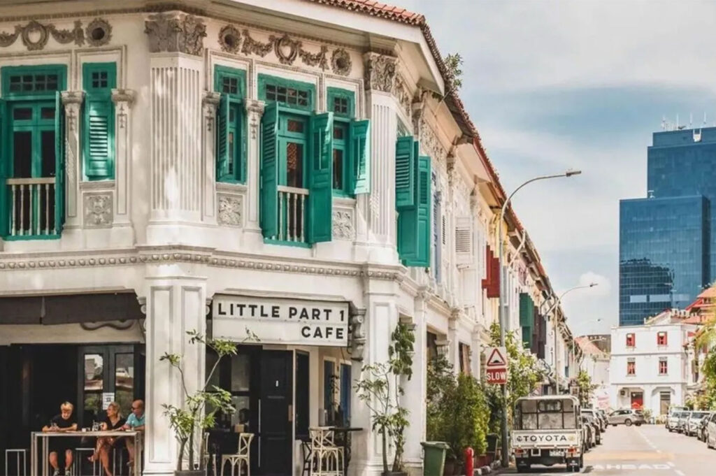 White heritage shophouse exterior with ornate columns and vibrant green shutters, featuring a ground-floor cafe sign reading "Little Part 1 Cafe," in a busy Singapore neighborhood.
