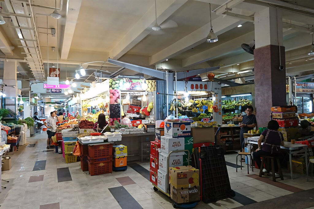 The brightly lit interior of a typical Singapore wet market (likely Tekka Centre), showing wide aisles, concrete floors, and numerous stalls selling fresh produce and packaged goods.