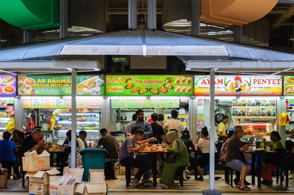 Bustling scene inside Tekka Centre hawker complex, showing customers eating at food stalls with brightly lit menu boards displaying Indian and Muslim cuisine.