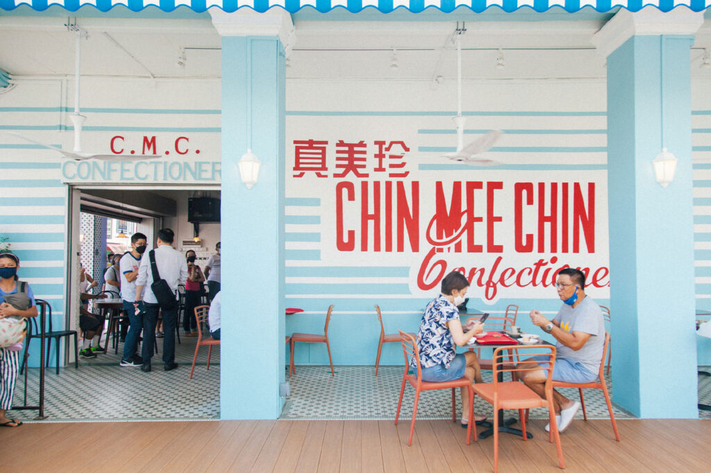 Exterior of the famous Chin Mee Chin Confectionery in Singapore, showing a retro-modern design with blue and white striped walls, red lettering, and customers seated at terracotta tables.