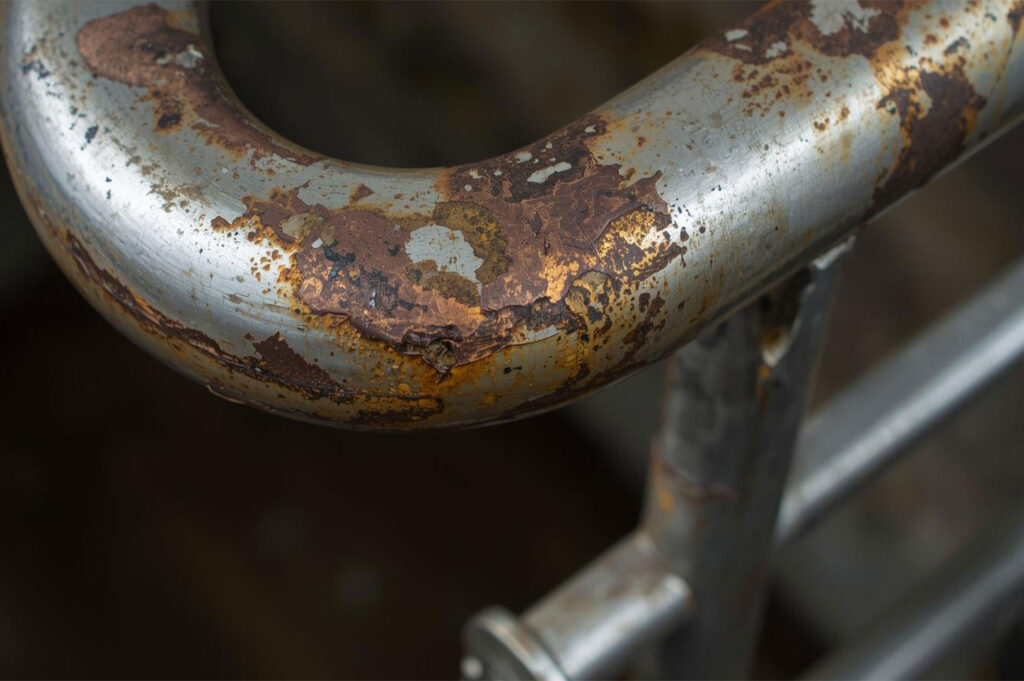 Extreme close-up of a metal railing or handrail, showing heavy oxidation, flaking paint, and large patches of dark brown and orange rust and corrosion.