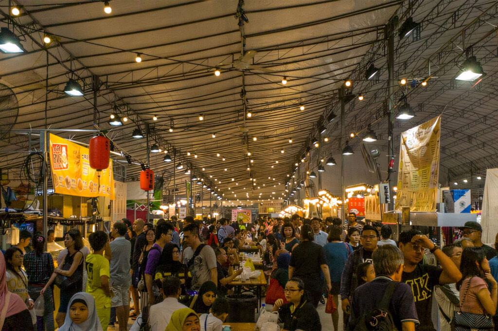 A densely crowded, low-light wide shot of a Pasar Malam (night market) in Southeast Asia, with strings of small lights overhead and numerous food stalls surrounded by shoppers and diners.