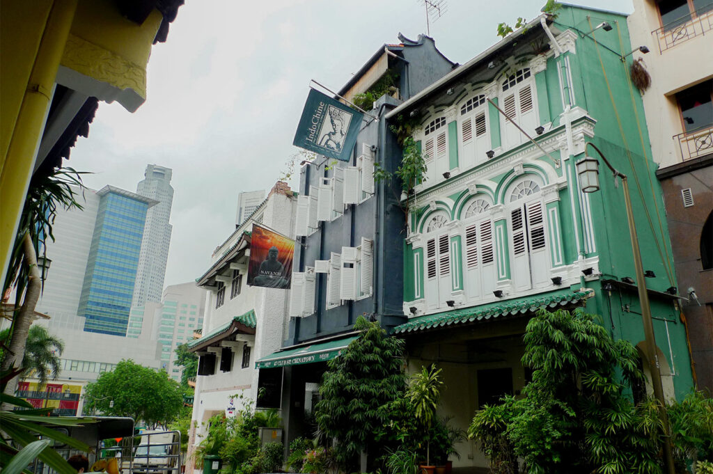 A street scene showing traditional Art Deco style shophouses in Singapore, featuring white shutters on a green and white facade, juxtaposed with modern skyscrapers in the background.