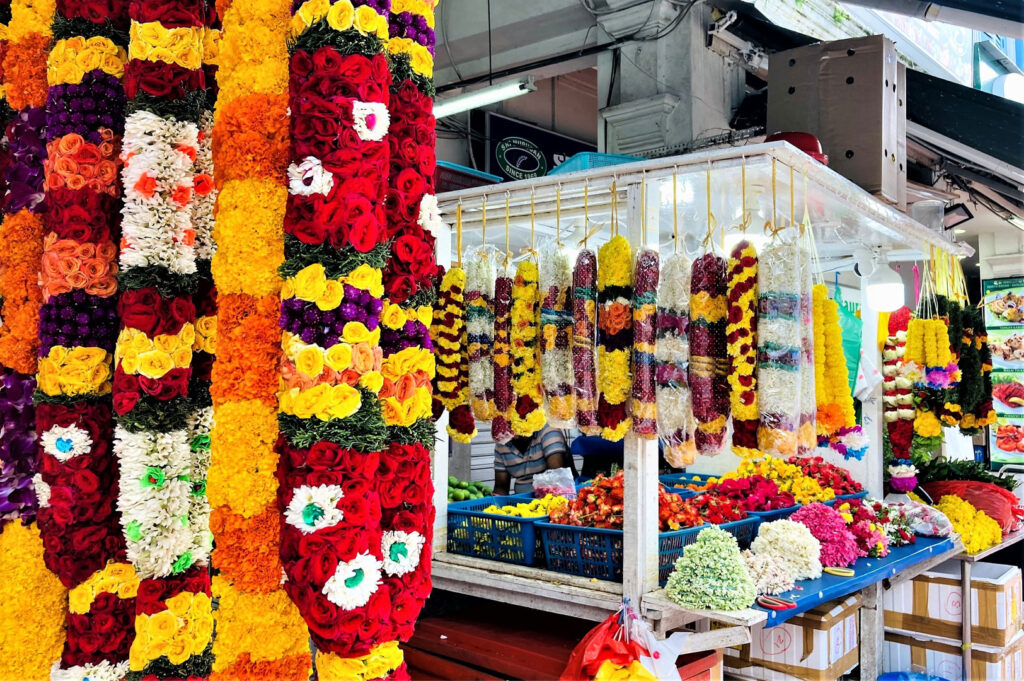 Close-up of a vibrant stall selling large, colorful Indian flower garlands (malai), made of marigolds and roses, hanging for sale at Tekka Centre.