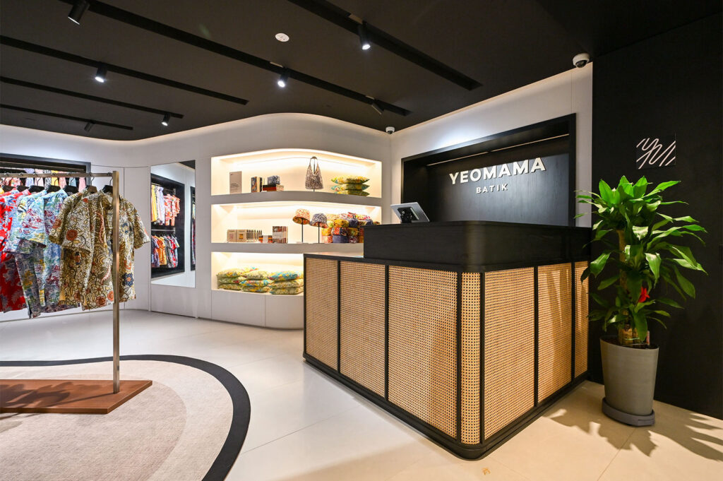 Interior of a modern retail boutique, "Yeomama Batik," featuring a rattan-paneled counter, minimalist shelves with accessories, and racks of colorful batik clothing.