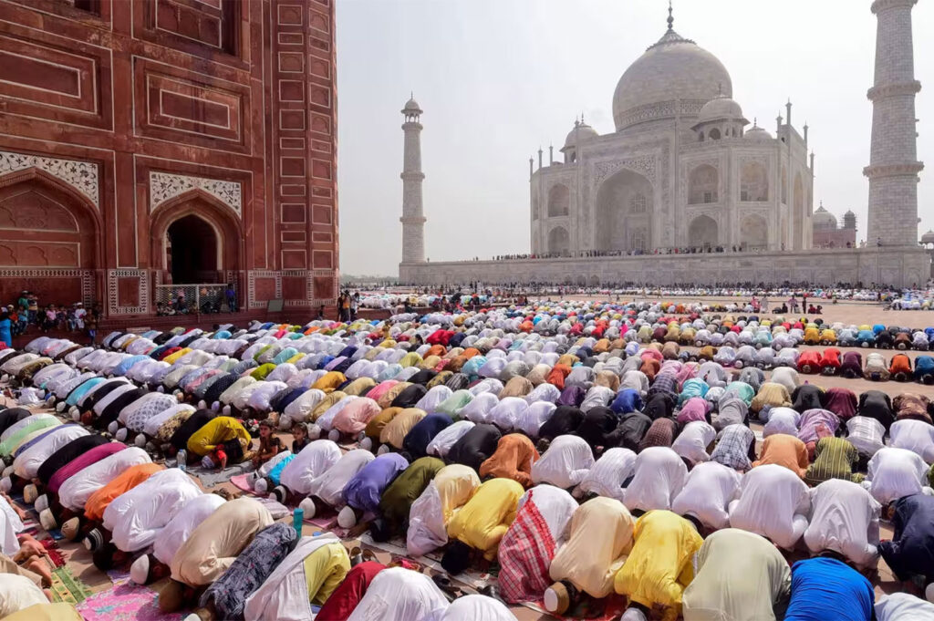 A vast crowd of Muslim worshippers of diverse colors praying in unison, likely for Hari Raya Puasa (Eid al-Fitr), kneeling outside a mosque or the Taj Mahal.