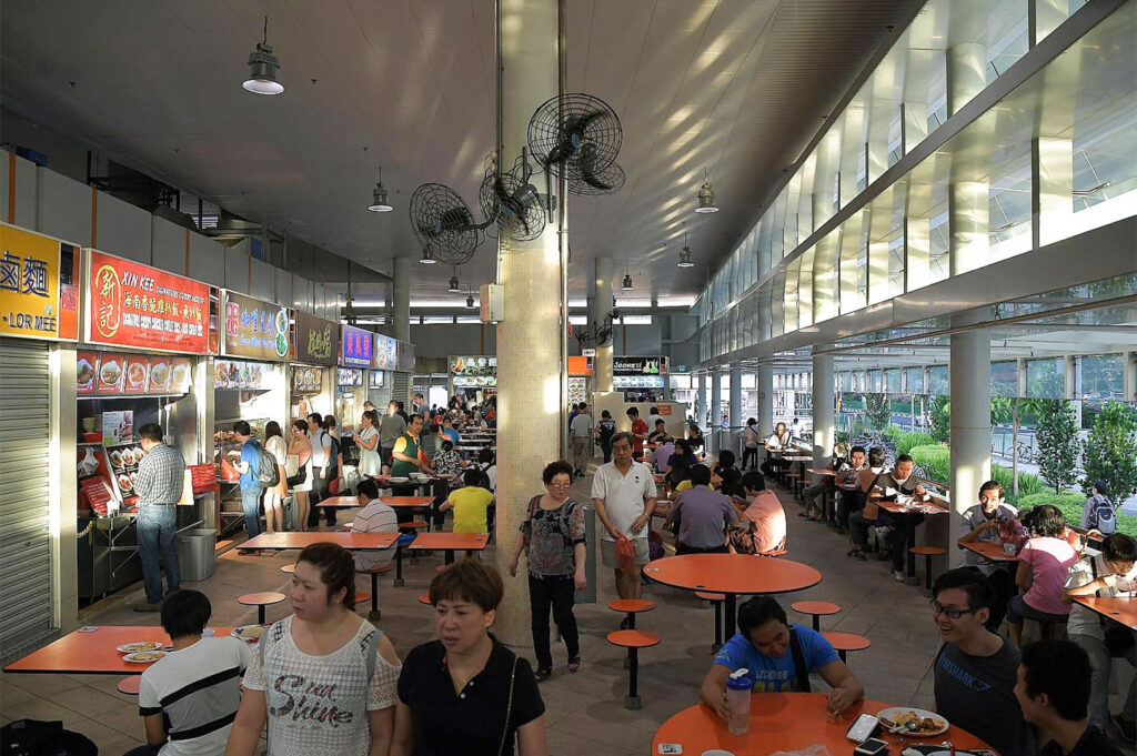 Wide interior view of a spacious, high-ceilinged Singapore hawker center during the day, filled with many patrons seated at round orange tables, with various stalls visible along the wall.