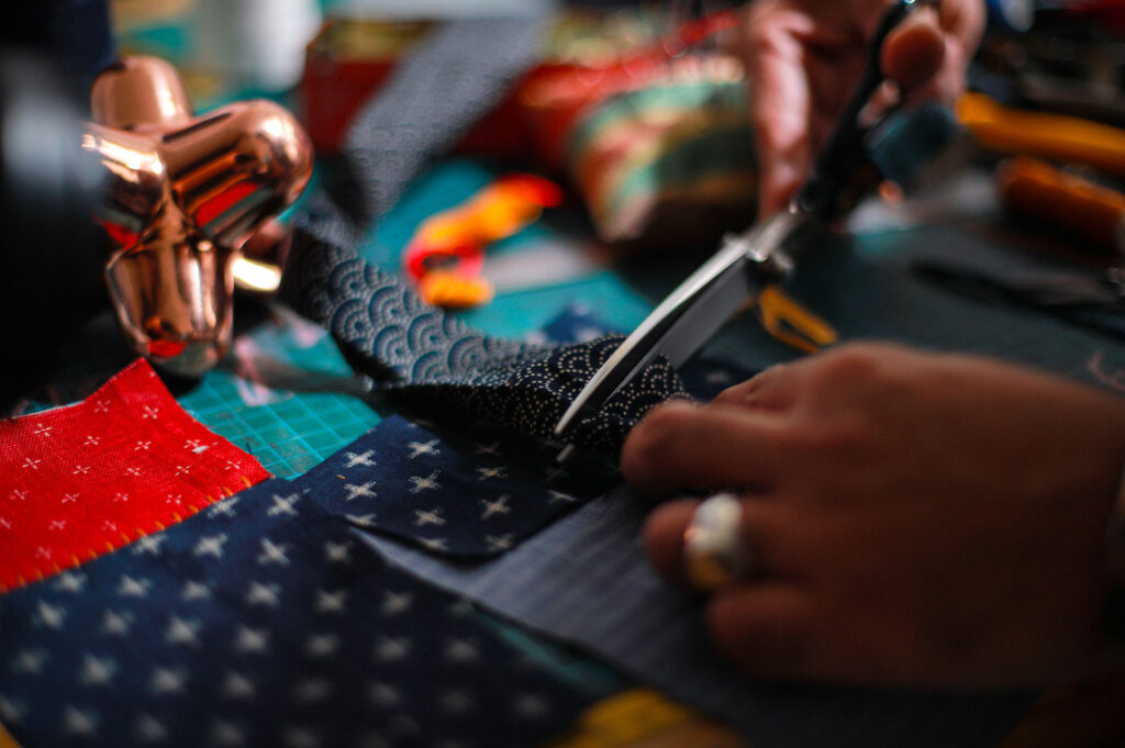 Close-up of a person's hands wearing a silver ring, carefully using scissors to cut a piece of patterned blue fabric on a colorful patchwork cutting mat.