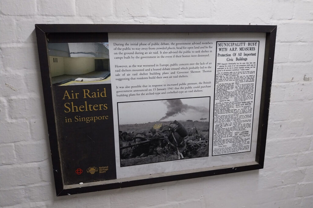 Informational plaque mounted on a white brick wall titled "Air Raid Shelters in Singapore," detailing the history of civil defense and public concerns during the initial phase of public debate, accompanied by historical text and a black-and-white photo of soldiers/shelter.