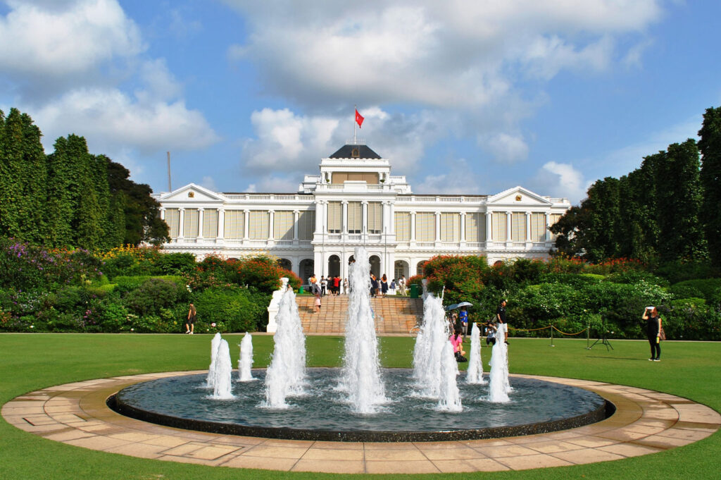 The grand, white colonial-style Istana, the official residence of the President of Singapore, viewed from a large green lawn with a circular fountain.