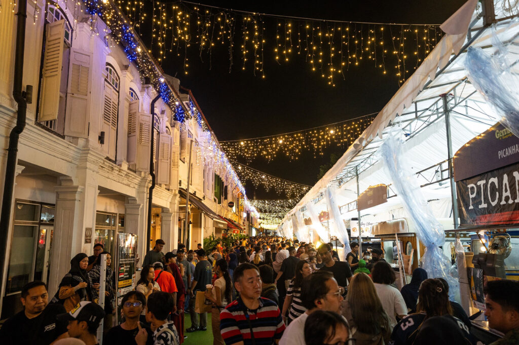 A vibrant night view of Kampong Gelam in Singapore, showing a temporary food bazaar or festival tent set up alongside historical, brightly lit shophouses with decorative string lights overhead.