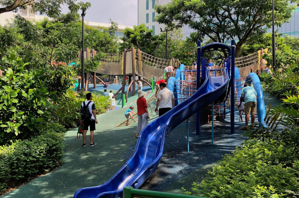 Children's playground nestled within a lush, tropical park setting, featuring a tall blue slide, extensive rope climbing nets, and surrounding greenery.