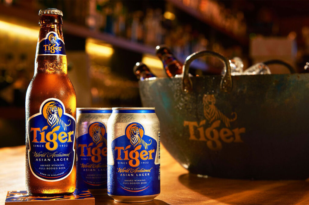 A promotional shot of a glass bottle and two cans of Tiger brand Asian Lager beer, with a branded bucket filled with ice and bottles visible in the background.