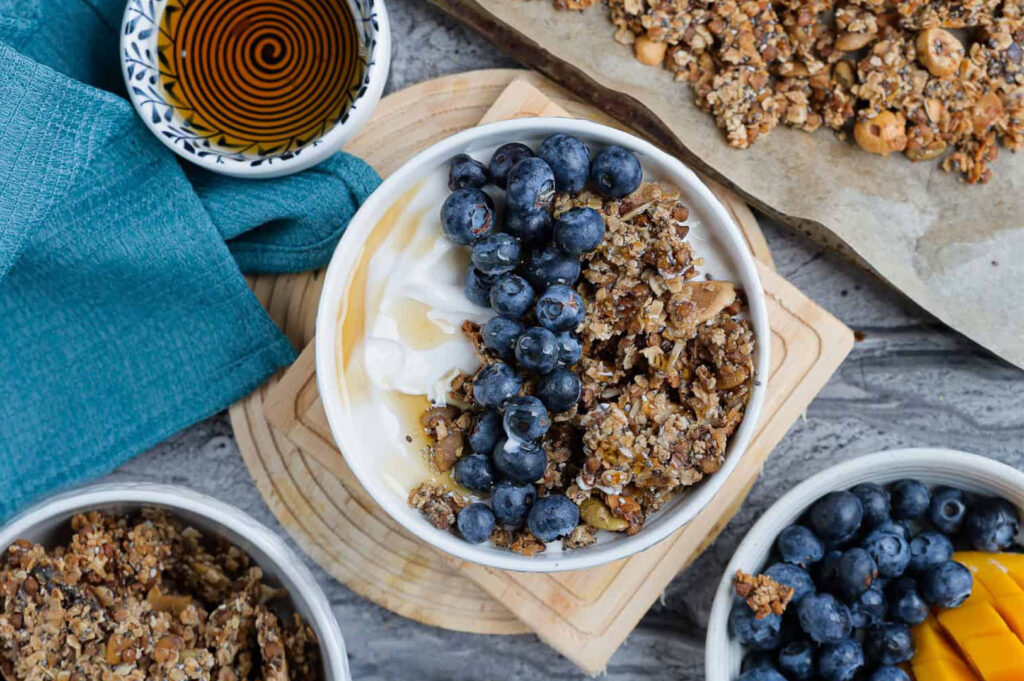 Overhead view of a breakfast bowl containing creamy yogurt, crunchy granola, fresh blueberries, and a drizzle of honey, with a bowl of honey and extra berries visible on the side.