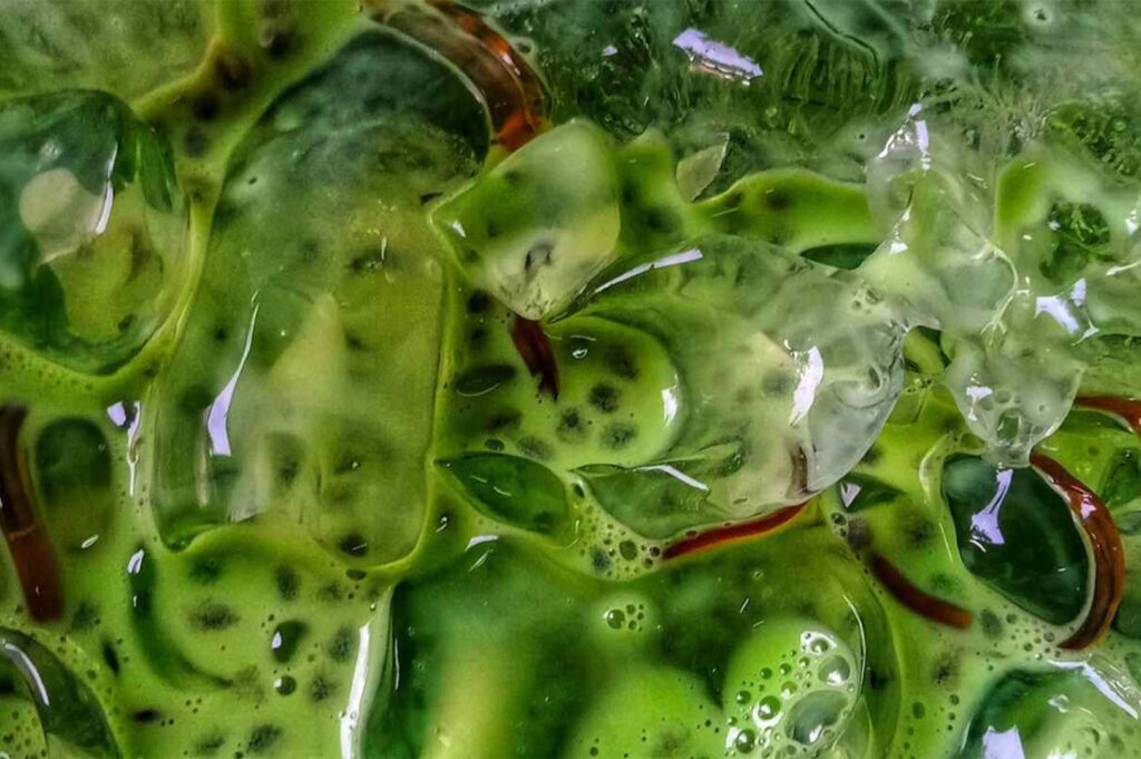 An extreme close-up, abstract macro shot of a vibrant green iced drink (likely Matcha) with condensation, ice cubes, and flecks of dark ingredients visible in the liquid.