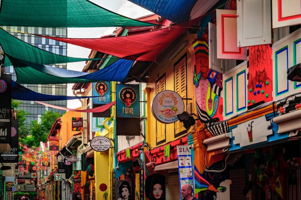 Vibrant street scene of colorful shophouses with art murals, advertising signs, and shade canopies over the pedestrian walk way in a cultural district.