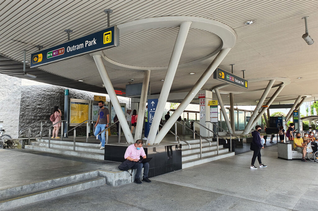 Exterior view of the modern Outram Park MRT Station exit in Singapore, with signage for multiple lines (EW16, NE3, TE17), and people entering and exiting the station.