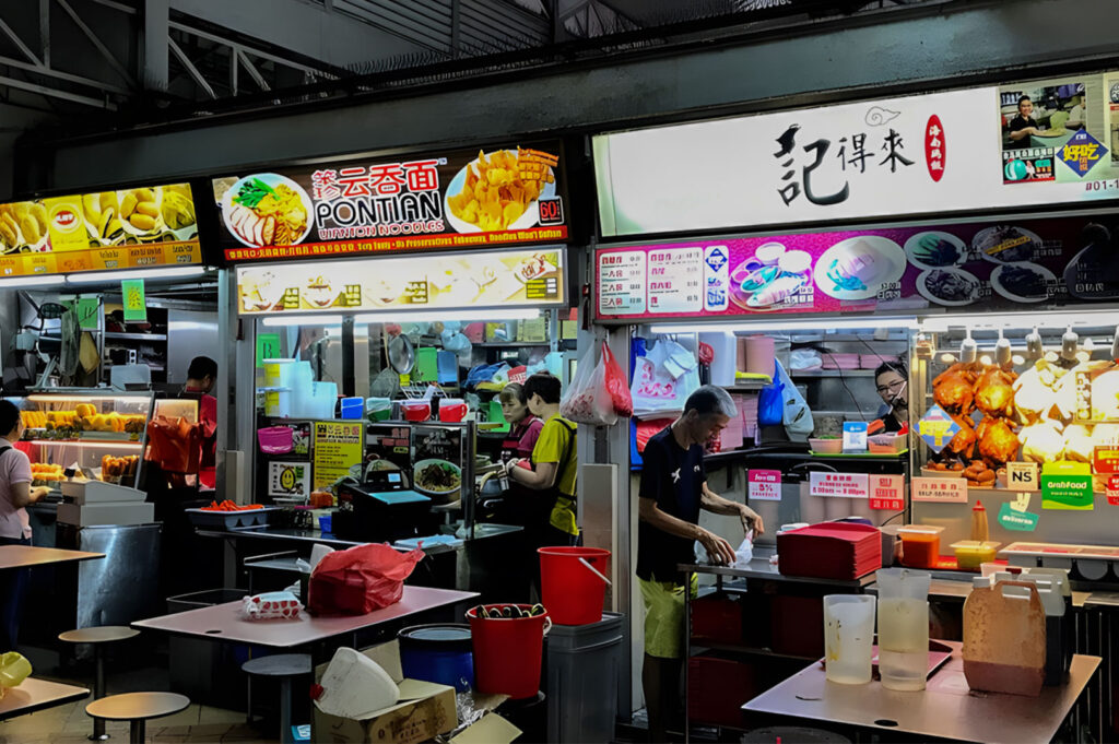 Close-up view of a vibrant hawker stall area at night, featuring brightly lit signs, including a stall specializing in "Pontian Noodles," with a vendor preparing food.
