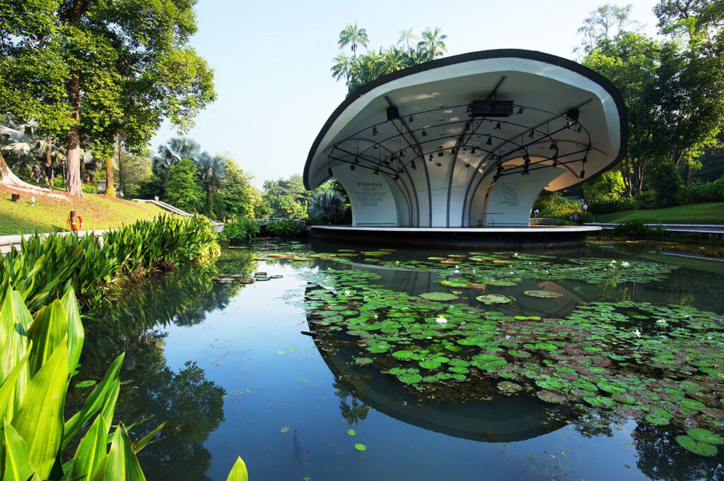The iconic, shell-shaped Shaw Foundation Symphony Stage (Bandstand) in the Singapore Botanic Gardens, reflected in a tranquil lake covered with lily pads.