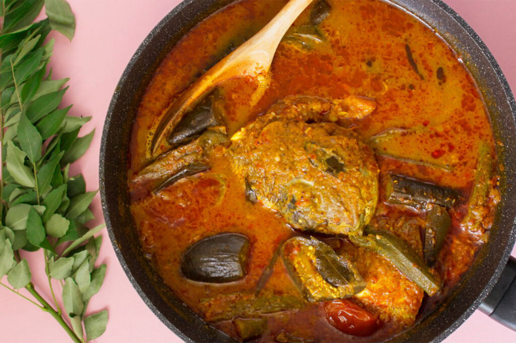 Overhead view of a black pan filled with a rich, reddish-orange Fish Head Curry, containing okra, eggplant, and curry leaves, resting on a pink surface.
