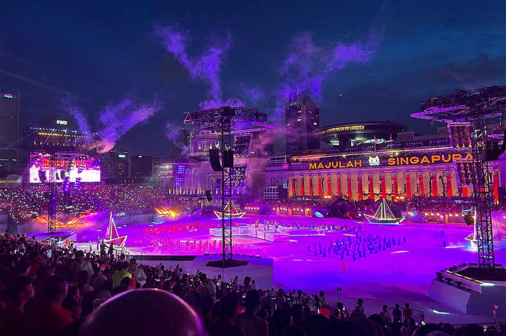 A massive, illuminated spectacle at the Singapore National Day Parade (NDP) at night, showing performers on the field and purple light effects over the city skyline.