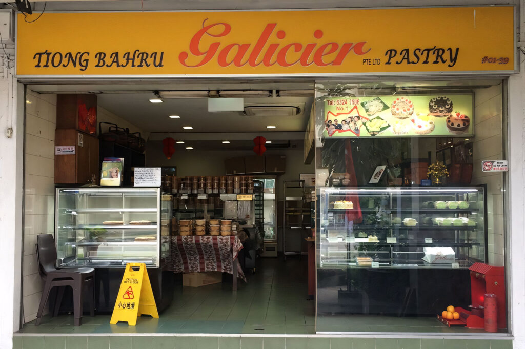 Storefront exterior of "Tiong Bahru Galicier Pastry," a traditional Singaporean bakery, with a large yellow sign and glass display cases showing traditional kueh and cakes.